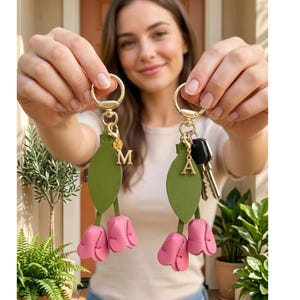 Könnte beinhalten: Zwei Schlüsselanhänger mit goldfarbener Hardware werden hochgehalten. Jeder Schlüsselanhänger hat ein grünes Blatt- und rosa Tulpen-Design. Einer hat den Buchstaben "M" und der andere den Buchstaben "A" angebracht. Schlüssel sind an dem Schlüsselanhänger mit dem Buchstaben "A" befestigt.