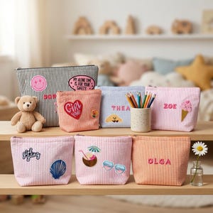 Könnte beinhalten: Eine Sammlung von gestreiften Stoffbeuteln in verschiedenen Farben mit gestickten Aufnähern und personalisierten Namen. Die Beutel zeigen Motive wie ein rosa Smiley-Gesicht, ein Herz, ein Croissant, eine Eistüte, eine Muschel, ein Kokosnussgetränk und herzförmige Sonnenbrillen.