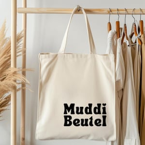 Könnte beinhalten: Cremefarbene Tragetasche mit den Worten "Muddi Beutel" in fetten, schwarzen Buchstaben. Die Tasche hat lange Henkel und hängt an einem Holzgestell mit anderen Kleidungsstücken. Der Hintergrund umfasst getrocknete Pflanzen.