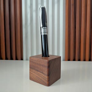 May include: A black pen with silver accents sits in a dark brown wooden pen holder.