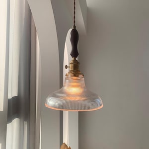 May include: A vintage-style pendant light with a clear, ribbed glass shade. The light fixture features a dark brown wooden handle, brass accents, and a brown cord. The light is turned on, emitting a warm glow.