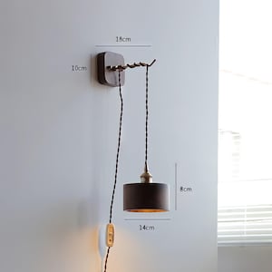 Op de afbeelding: Een wandlamp met een donkerbruine kap, 14 cm in diameter en 8 cm hoog, hangt aan een decoratieve armatuur. De armatuur is bevestigd aan een donkerbruine houten basis, 18 cm breed en 10 cm hoog, met een gedraaid snoer en een aan/uit-schakelaar.