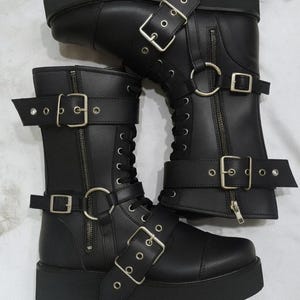May include: Pair of black platform boots with multiple straps, buckles, and zippers. The boots have a chunky sole and a mid-calf height. The design features silver-toned hardware and a lace-up front. The boots are made of a smooth, matte material.