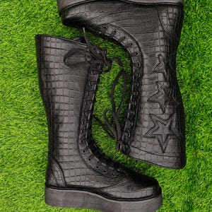 May include: A pair of black knee-high boots with a crocodile-embossed texture, featuring a thick platform sole, black laces, and three stitched star designs on the outer side of each boot shaft. The boots have a rounded toe and a tall silhouette.