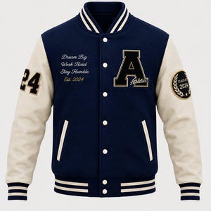 May include: A navy blue varsity jacket with cream-colored sleeves, featuring a large letter A with the word Athletics, the text Dream Big Work Hard Stay Humble Est. 2024, the number 24 on the right sleeve, and a Class of 2024 patch on the left sleeve.