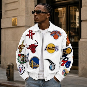 Może przedstawiać: Biała kurtka typu varsity z różnymi wyhaftowanymi logotypami drużyn NBA oraz naszywkami o tematyce koszykarskiej na przodzie i rękawach. Kurtka posiada klasyczny kołnierz, zapięcia na zatrzaski oraz ściągacze przy mankietach.