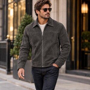 K&ouml;nnte beinhalten: Eine anthrazitfarbene Wildlederjacke mit klassischem Kragen, durchgehendem silberfarbenem Metallrei&szlig;verschluss und langen &Auml;rmeln. Die Jacke wird &uuml;ber einem schlichten wei&szlig;en Rundhals-Shirt getragen und mit einer dunkelblauen Jeans kombiniert. Sie hat eine horizontale Naht &uuml;ber der Brust und zwei Seitentaschen.