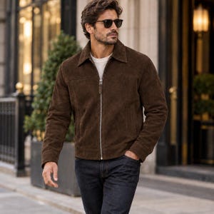 May include: A dark brown suede jacket with a classic collar, long sleeves, and a full-length silver-toned metal zipper closure. The jacket features a horizontal seam across the chest and is worn over a white crew-neck shirt and dark denim jeans.
