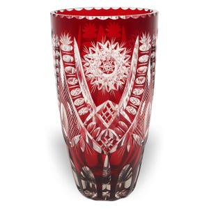 Op de afbeelding: Een hoge, cilindrische vaas van dieprood glas met ingewikkelde, helder geslepen geometrische patronen. Het ontwerp bevat sterren, kruisarceringen en ovale vormen die in het oppervlak zijn geslepen en het heldere glas onder het rood onthullen.