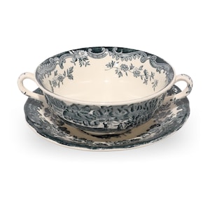 Może przedstawiać: Biało-niebieska ceramiczna miska do zupy z dwoma uchwytami, spoczywająca na pasującym spodku. Miska i spodek mają szczegółową scenę krajobrazu i kwiatowy wzór obramowania. Zestaw jest idealny do serwowania zupy.