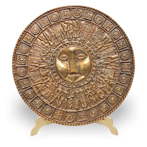 May include: A bronze-colored decorative plate with a sun face design at the center. The plate features intricate relief patterns of figures and geometric shapes. It is displayed on a gold-colored stand, showcasing its artistic detail.