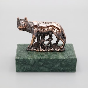 May include: A bronze sculpture of the Capitoline Wolf nursing Romulus and Remus, mounted on a rectangular green marble base. The wolf is detailed with a textured coat and stands over the twins.