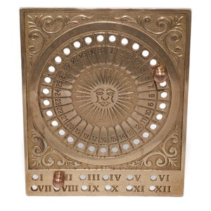 Può includere: Un calendario vintage in ottone con un disegno a forma di sole al centro. Il calendario ha un quadrante circolare con i numeri da 1 a 31 e una piccola sfera che indica la data. La base presenta numeri romani I-XII. Il calendario è quadrato e presenta decorazioni.