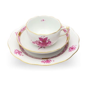May include: A white porcelain teacup, saucer, and plate set. The set features a pink floral design with gold trim. The teacup has a scalloped rim, and the plate and saucer have a similar design. A classic tea set.