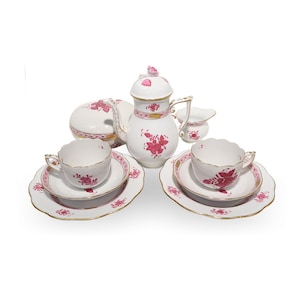 May include: A white porcelain tea set with a pink floral design. The set includes a teapot, sugar bowl, creamer, two teacups, and saucers. The rims are trimmed with gold. The teacups and saucers have a scalloped edge.