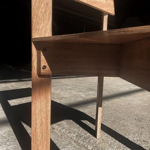May include: Close-up of a wooden chair with a light brown finish. The chair has a simple design with a rectangular seat and back. The chair is made of plywood and has a natural wood grain.