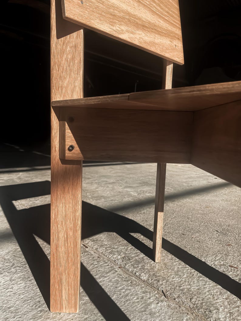 May include: Close-up of a wooden chair with a light brown finish. The chair has a simple design with a rectangular seat and back. The chair is made of plywood and has a natural wood grain.
