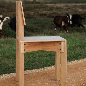 May include: A wooden chair with a unique design, featuring a tall back and a simple, rectangular seat. The chair is made of light-colored wood and has a natural finish. It is standing on a dirt path with a grassy field in the background.