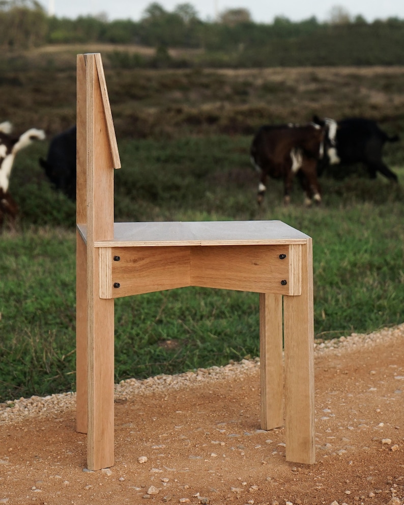May include: A wooden chair with a unique design, featuring a tall back and a simple, rectangular seat. The chair is made of light-colored wood and has a natural finish. It is standing on a dirt path with a grassy field in the background.