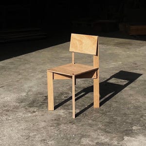 May include: A simple wooden chair with a light brown finish. The chair has a rectangular seat and a back that is slightly angled. The chair is made of plywood and has a minimalist design.