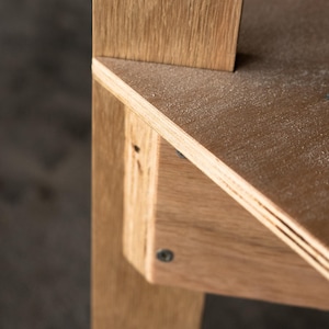 May include: Close-up of a wooden shelf unit with a light wood finish. The shelf is made of plywood and has a slightly angled edge. The shelf is attached to a vertical support beam with a screw.