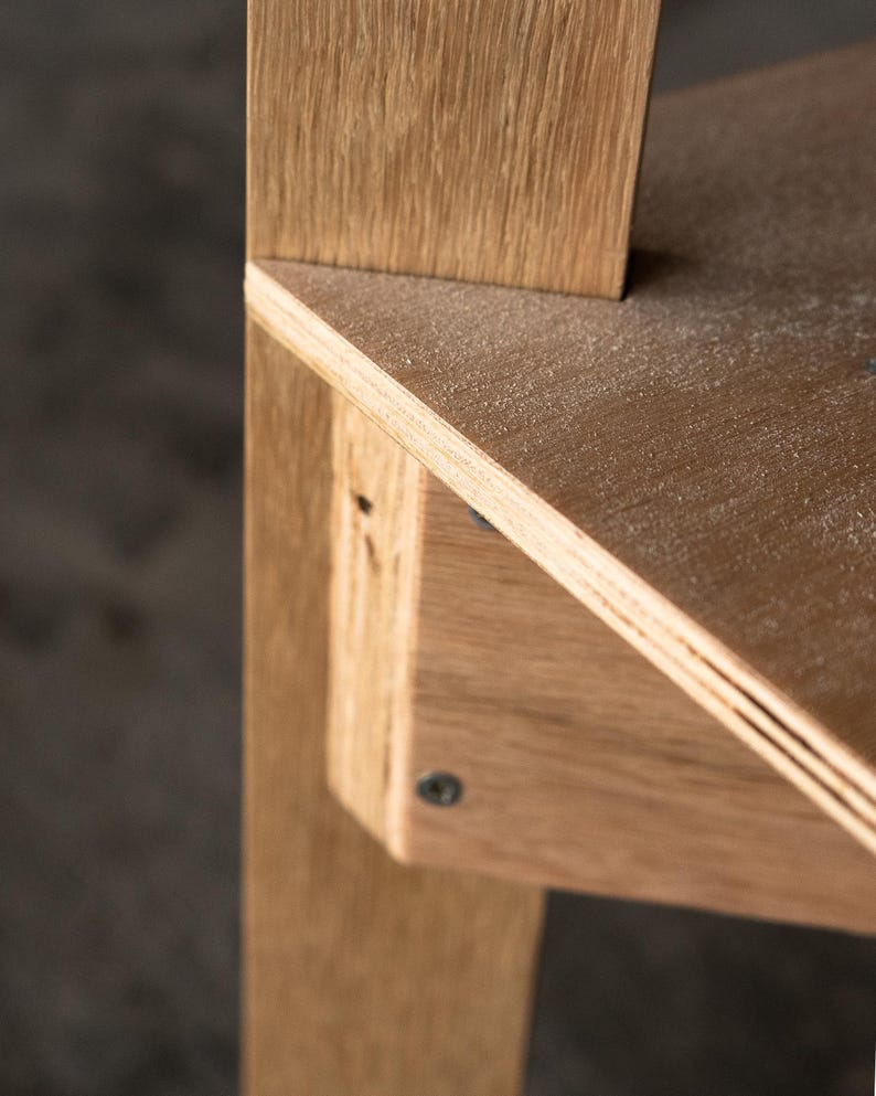 May include: Close-up of a wooden shelf unit with a light wood finish. The shelf is made of plywood and has a slightly angled edge. The shelf is attached to a vertical support beam with a screw.