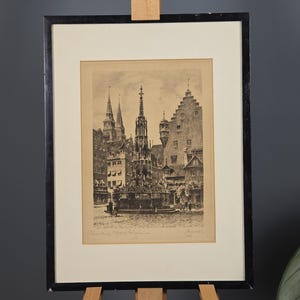 May include: A framed black and white etching depicting a town square with a central gothic fountain, surrounded by historic buildings with gabled roofs and spires. The artwork is mounted in a cream-coloured mat and a simple black frame.