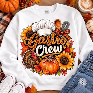 Peut inclure: Un sweat-shirt blanc à col rond avec une impression graphique portant le texte Gastro Crew. Le motif comprend une toque de chef, une cuillère en bois, un fouet, une dinde, une citrouille, des tournesols et des feuilles d'automne.