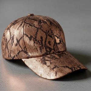May include: A baseball cap with a snakeskin pattern in bronze and black. The cap has a curved peak and a button on top. The hat is made of a textured material.