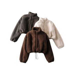 May include: Three cropped fleece jackets in brown, dark grey, and cream. Each jacket features a high collar and a full-length zip. The brown jacket in the foreground has a drawstring at the hem. The jackets are made of a soft, textured material.