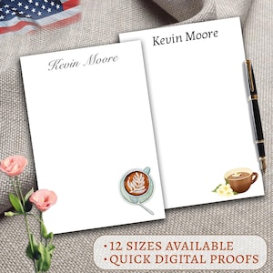 May include: Two white notepads with the name "Kevin Moore" printed at the top. One notepad features a coffee cup illustration with a leaf design, and the other has a coffee cup with flowers. A black pen rests on the side.