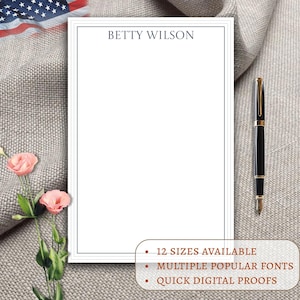 May include: A white notepad with a blue border and the name "BETTY WILSON" printed at the top. A black and gold pen rests on the right side. The notepad is on a textured gray surface. Text at the bottom reads: "12 SIZES AVAILABLE", "MULTIPLE POPULAR FONTS", "QUICK DIGITAL PROOFS".
