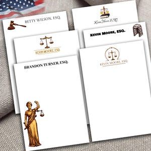 May include: Four white notepads with different legal-themed designs. The top notepad has a gavel and the text "BETTY WILSON, ESQ.". The second notepad has a scale of justice and the text "KEVIN MOORE, ESQ.". The third notepad has a scale of justice and the text "KEVIN MOORE, ESQ.". The fourth notepad has a scale of justice and the text "KEVIN MOORE, ESQ.". The bottom notepad has a drawing of the goddess of justice and the text "BRANDON TURNER, ESQ."