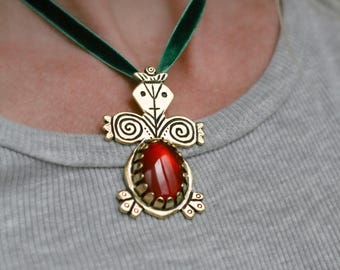 May include: A gold-toned pendant necklace with a red gemstone center and an ornate design. The pendant is suspended from a dark green velvet ribbon. The necklace is worn against a gray shirt.