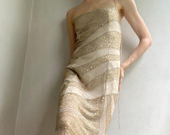 May include: A floor-length, cream-colored dress with gold sequin stripes. The dress features thin spaghetti straps and a fitted bodice that flows into a slightly flared skirt. The gold sequins create a shimmering effect, adding a touch of glamour to the design.