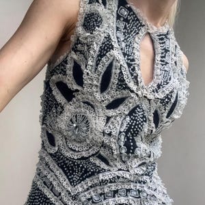May include: A sleeveless black top with intricate white lace and sequin embellishments. The top features a keyhole neckline and a flared, cream-colored skirt. The design includes floral and geometric patterns, creating a vintage or retro aesthetic.
