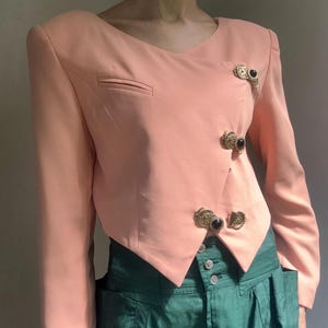 May include: A peach-colored cropped blazer with long sleeves, a V-neckline, and an asymmetrical front closure. The jacket features three decorative gold-toned buttons with black circular centers. It is paired with high-waisted green trousers.