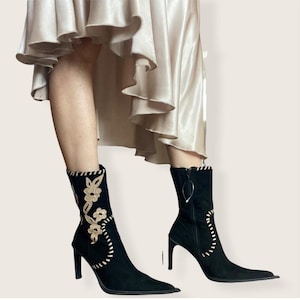 May include: Black suede ankle boots with pointed toes and stiletto heels. The boots feature beige floral embroidery and decorative stitching. A zipper runs up the side of one boot. The boots are worn with a cream-colored skirt.