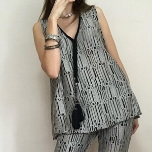 May include: Silver sleeveless top and matching trousers set. The outfit has a vertical striped pattern with black accents. A black tassel hangs from the V-neckline of the top. The fabric has a shimmering, textured appearance, creating a glamorous look.