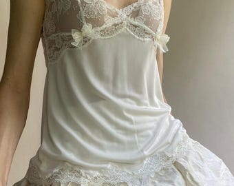 Può includere: Una canotta bianca con corpetto in pizzo trasparente, scollo a V, spalline sottili e due piccoli fiocchi decorativi sul petto. La parte inferiore del capo è realizzata in tessuto liscio e opaco con un bordo in pizzo sull'orlo.