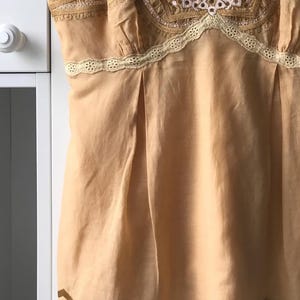 May include: A peach-colored camisole with delicate floral embroidery and lace trim. The top features a V-neckline and a pleated design, with floral embroidery along the straps and hem. The camisole is made of a lightweight fabric.