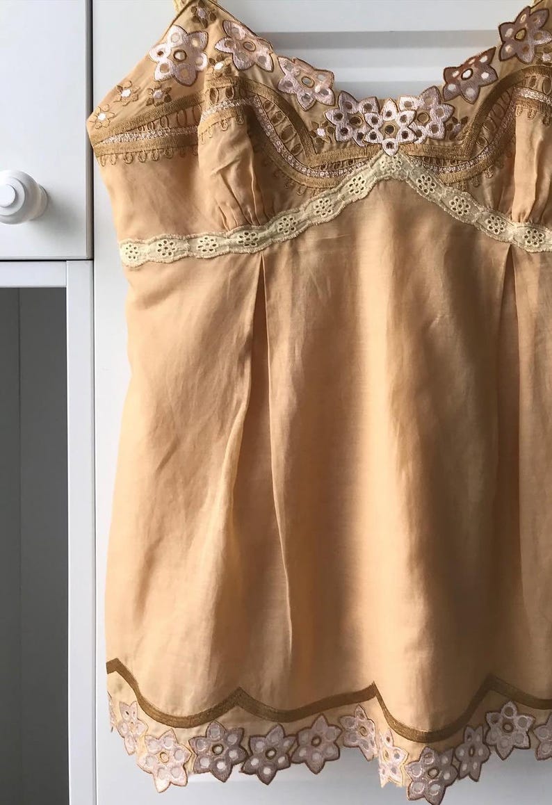 May include: A peach-colored camisole with delicate floral embroidery and lace trim. The top features a V-neckline and a pleated design, with floral embroidery along the straps and hem. The camisole is made of a lightweight fabric.