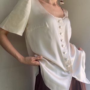 May include: A cream-colored blouse with short sleeves, a scoop neckline, and a vertical row of matching fabric-covered buttons down the front. The garment features a smooth, satin-like finish and a slightly asymmetrical hemline that drapes at the hip.