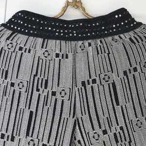 May include: Close-up of silver and black patterned trousers. The trousers have a black sequined waistband. The fabric has a textured appearance with vertical stripes and geometric shapes.