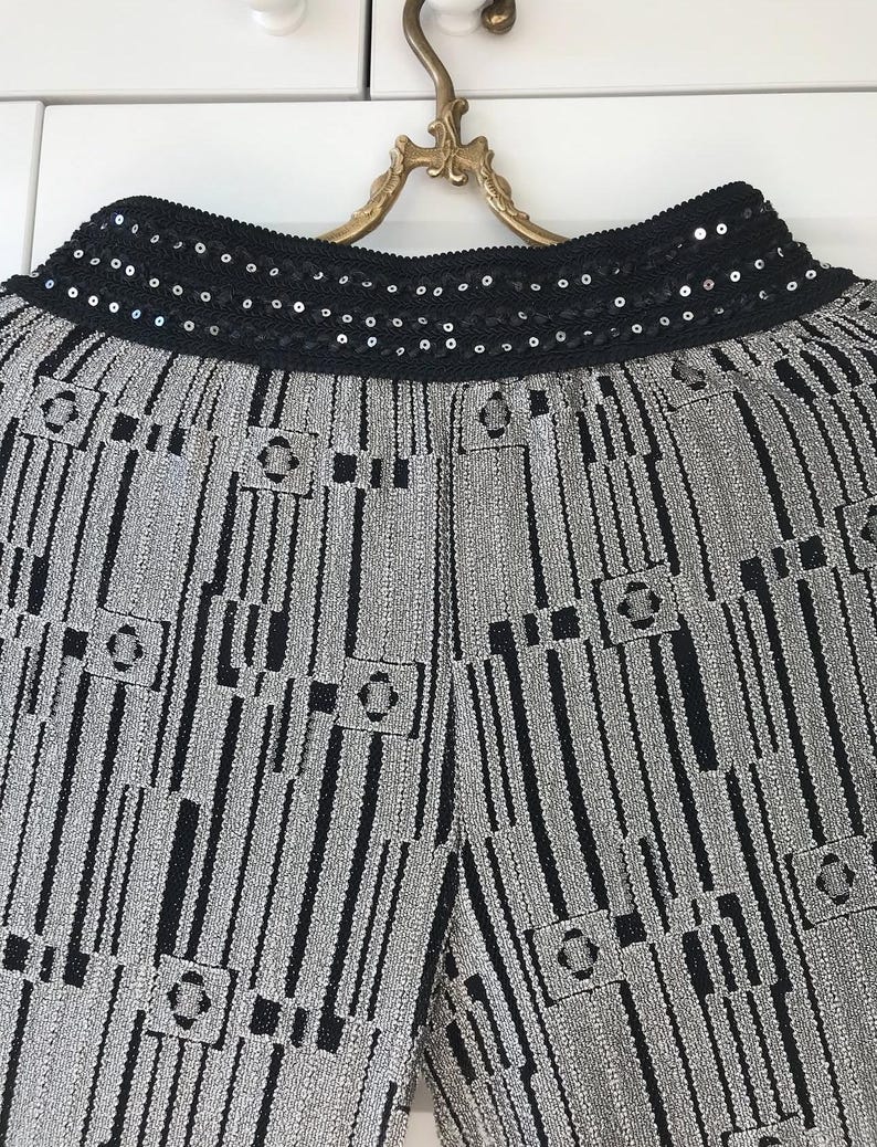 May include: Close-up of silver and black patterned trousers. The trousers have a black sequined waistband. The fabric has a textured appearance with vertical stripes and geometric shapes.