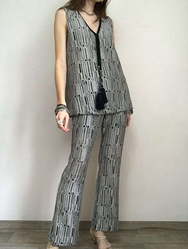 May include: A two-piece outfit comprising a sleeveless vest and flared trousers. The outfit is patterned with vertical black and silver stripes and geometric shapes. The vest has a V-neck with black trim and a tassel. The trousers are high-waisted.