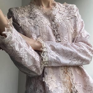 以下が含まれることがあります： 長袖とハイカラーの薄いピンク色のレースジャケット。 ジャケットは、袖と襟にスカラップの縁取りが施された、複雑な花のレースのディテールが特徴です。 デリケートでフェミニンな衣服です。