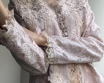 May include: A light pink lace jacket with long sleeves and a high collar. The jacket features intricate floral lace detailing throughout, with scalloped edges on the sleeves and collar. The jacket is a delicate, feminine garment.