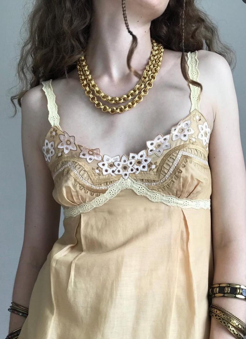 May include: A light yellow camisole top with floral embroidery and lace trim. A gold chain necklace and multiple gold bracelets are also visible. The top has thin straps and a scoop neckline.