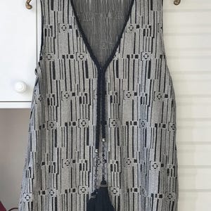 May include: Sleeveless vest featuring a geometric design in black and grey. The vest has a V-neckline trimmed with black fabric and a black braided tie with beaded accents and a tassel. The garment is suspended on a gold-coloured hanger.
